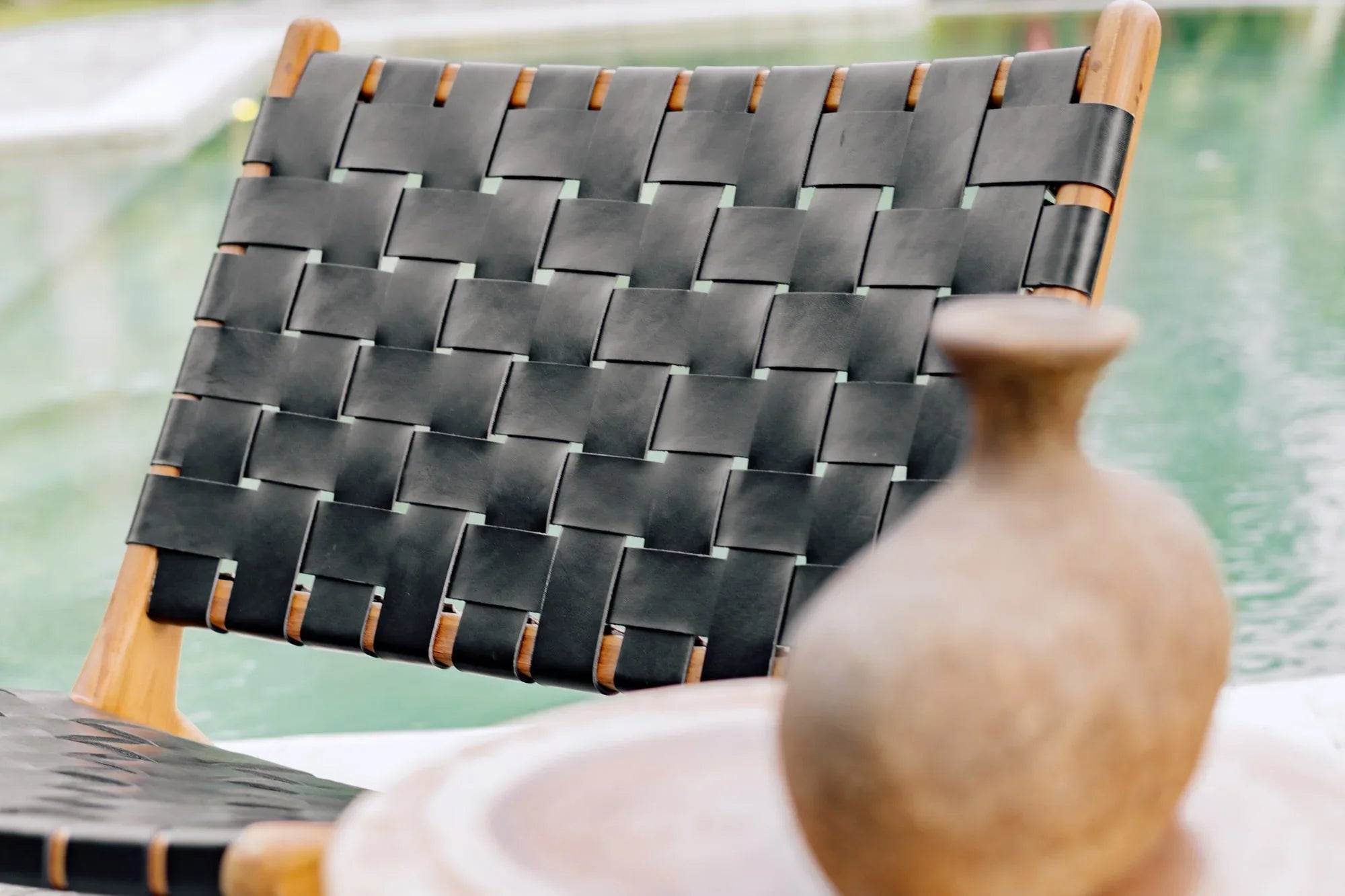Handmade woven black leather and wood chair by a pool, rustic vase in foreground, Kayu Living
