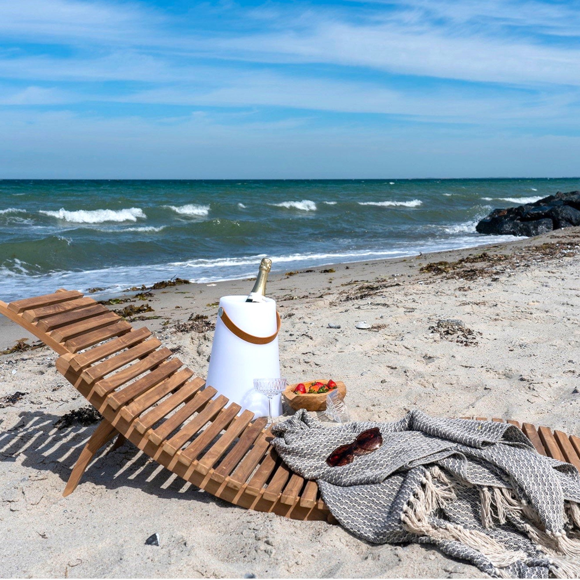 Kayu Living Arrieta natural teak sun lounger on a sandy beach with champagne and strawberries.