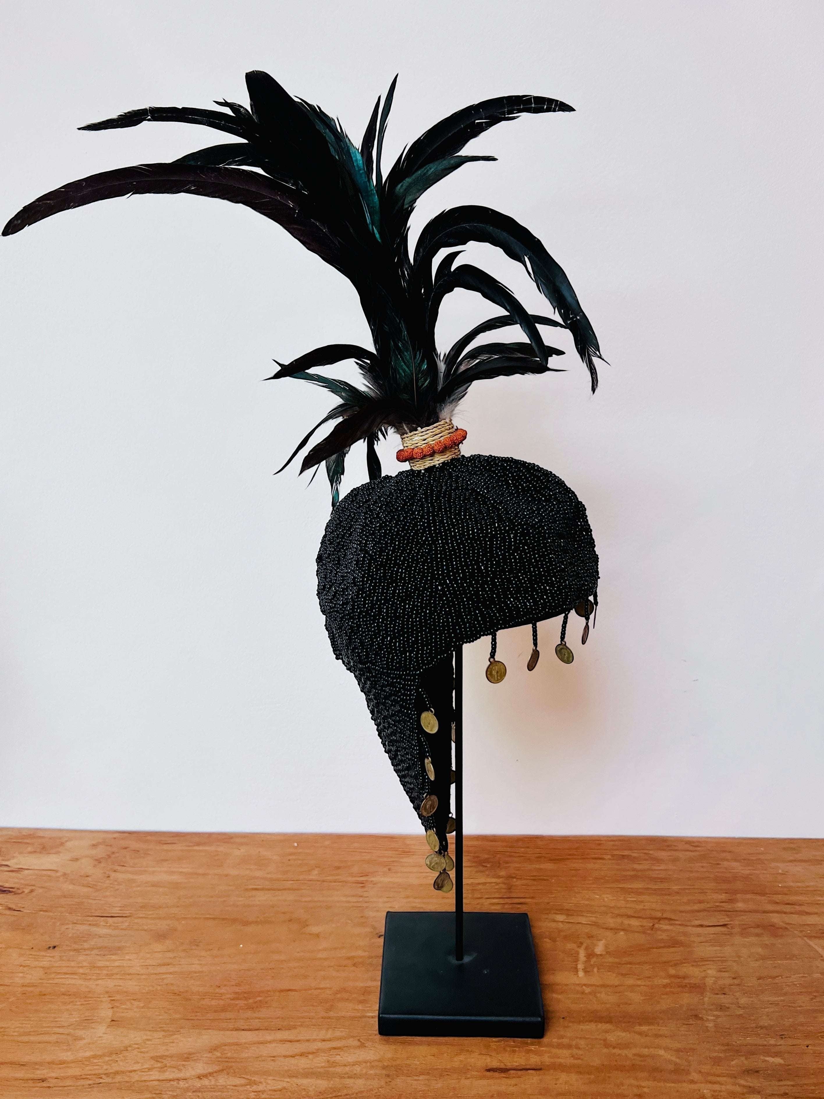 Kayu Living black beaded hat with feathers and coin accents, displayed on a stand. Handmade decorative art piece.