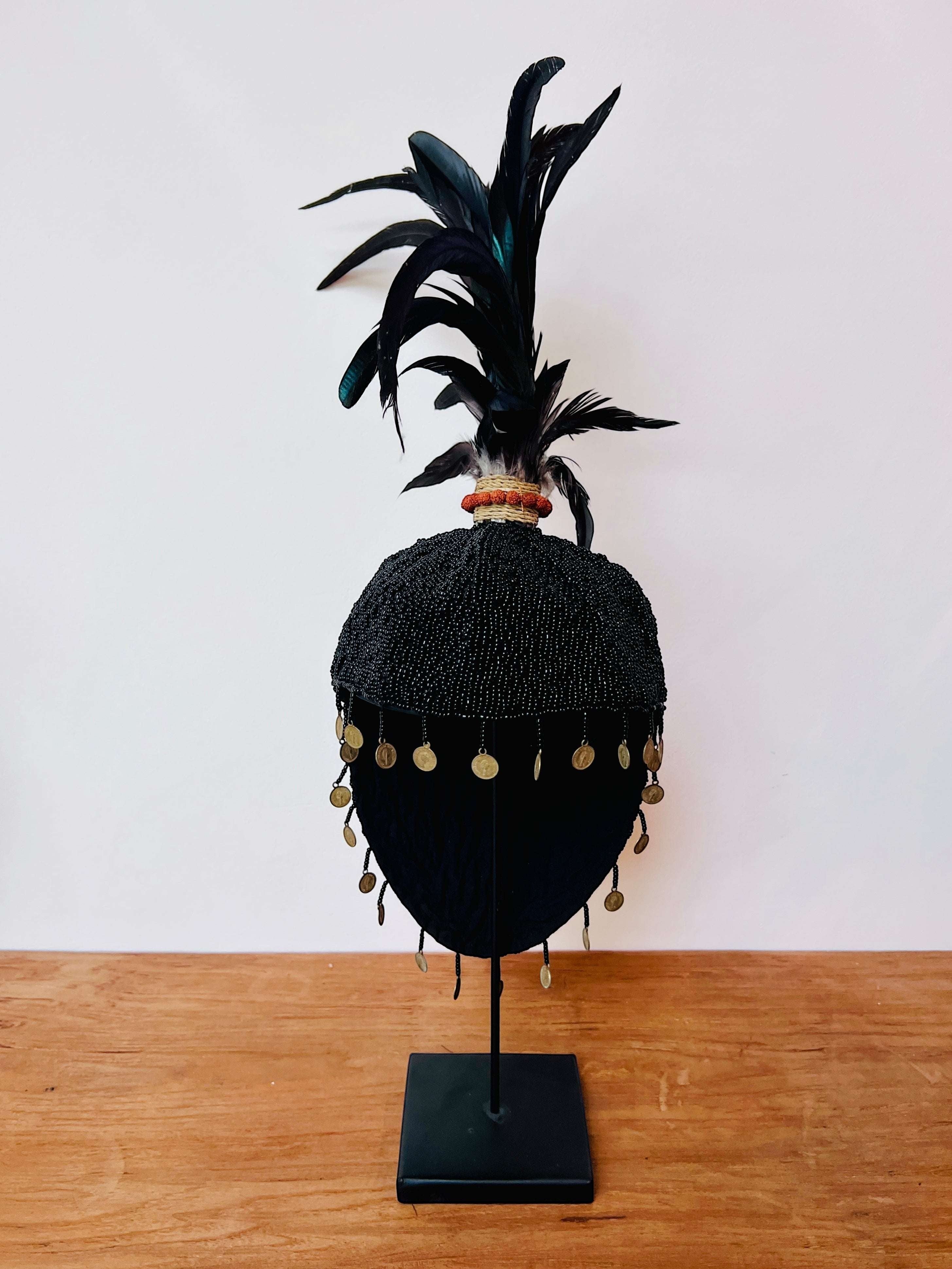 Kayu Living black beaded hat decoration with feathers, seeds, and coins on a stand.