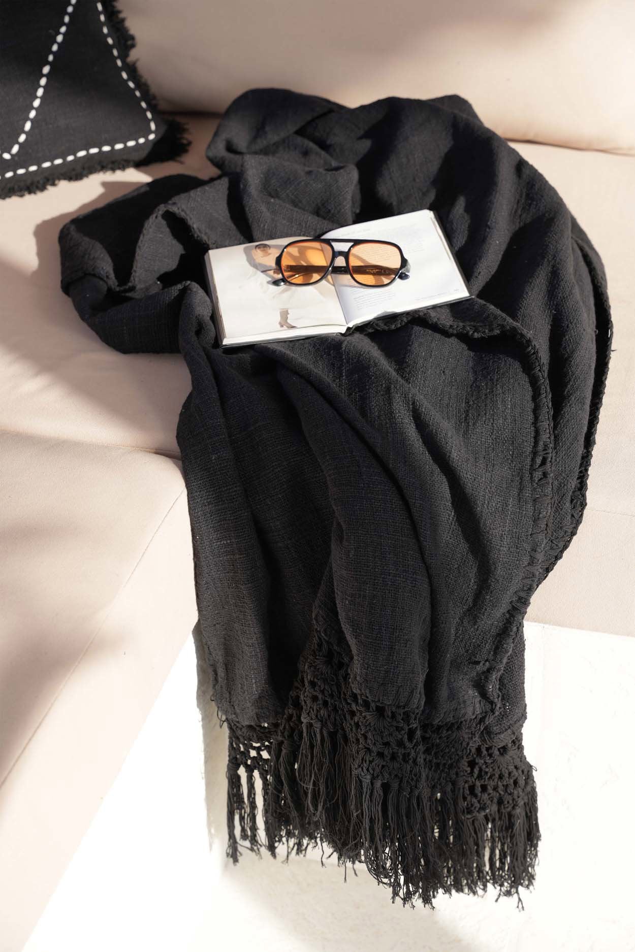 Kayu Living black cotton Gypsy bed throw with bohemian fringe detail draped over a couch with a book and sunglasses.