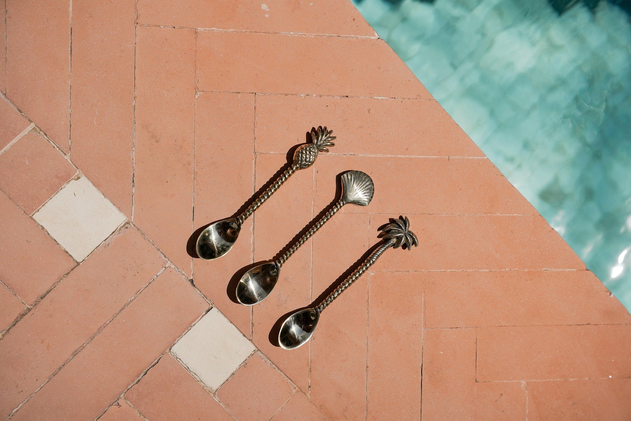 Kayu Living handcrafted brass spoons with pineapple, shell, and palm tree motifs on a terracotta tiled surface.