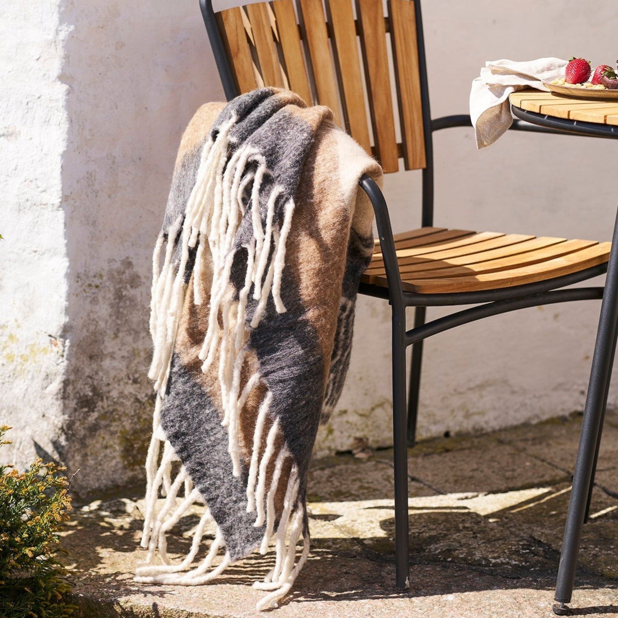 Kayu Living Carmel plaid wool blend throw in black and brown with fringe detail draped over a wooden patio chair.