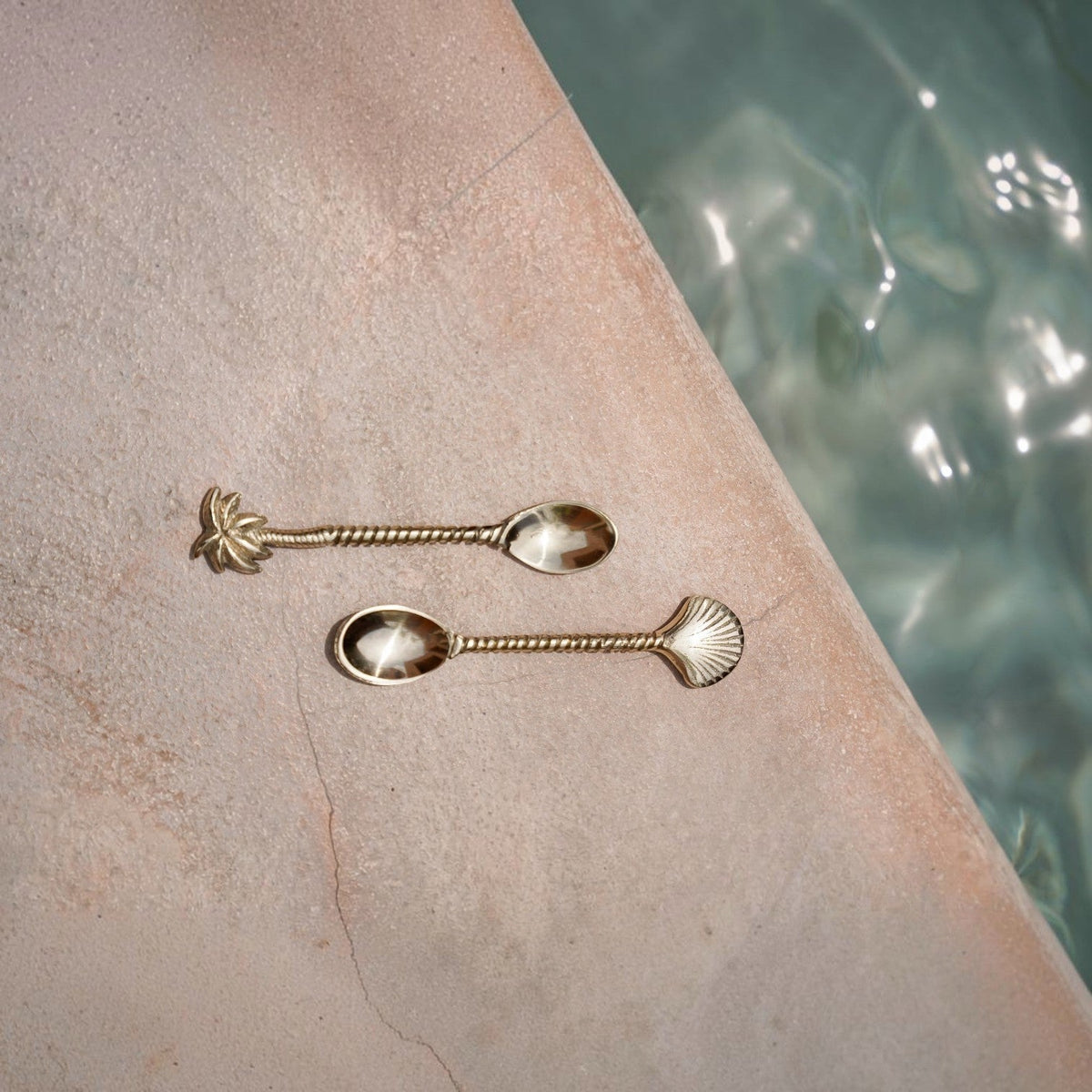 Kayu Living gold brass spoons with palm tree and shell motifs on a stone surface near water