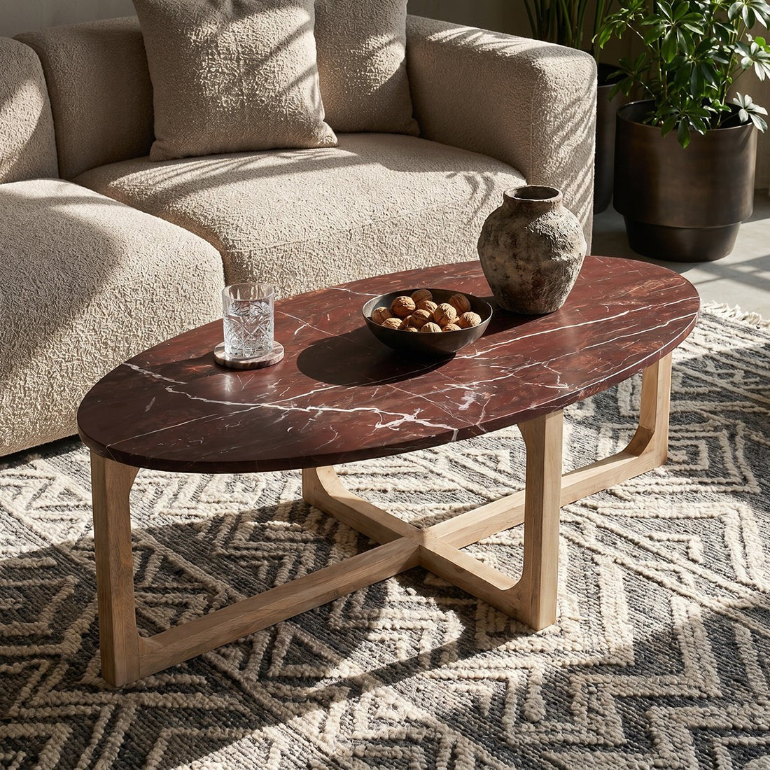 Kayu Living oval marble coffee table with bordeaux top and white veining on a natural mango wood base. Features a glass of water, bowl of nuts, and vase.