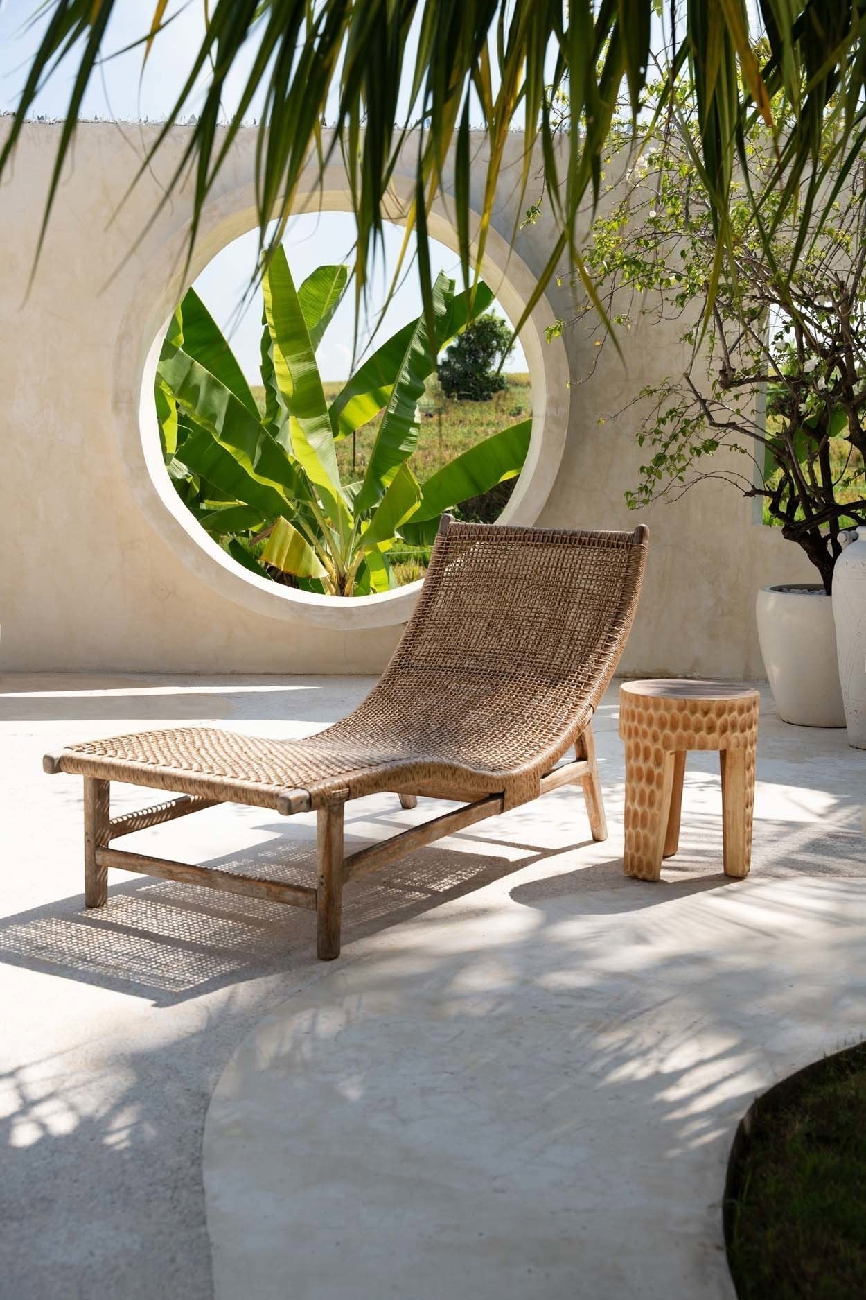 Kayu Living Paramo Stool in natural suar wood with carved texture, placed next to a woven lounge chair on a patio.