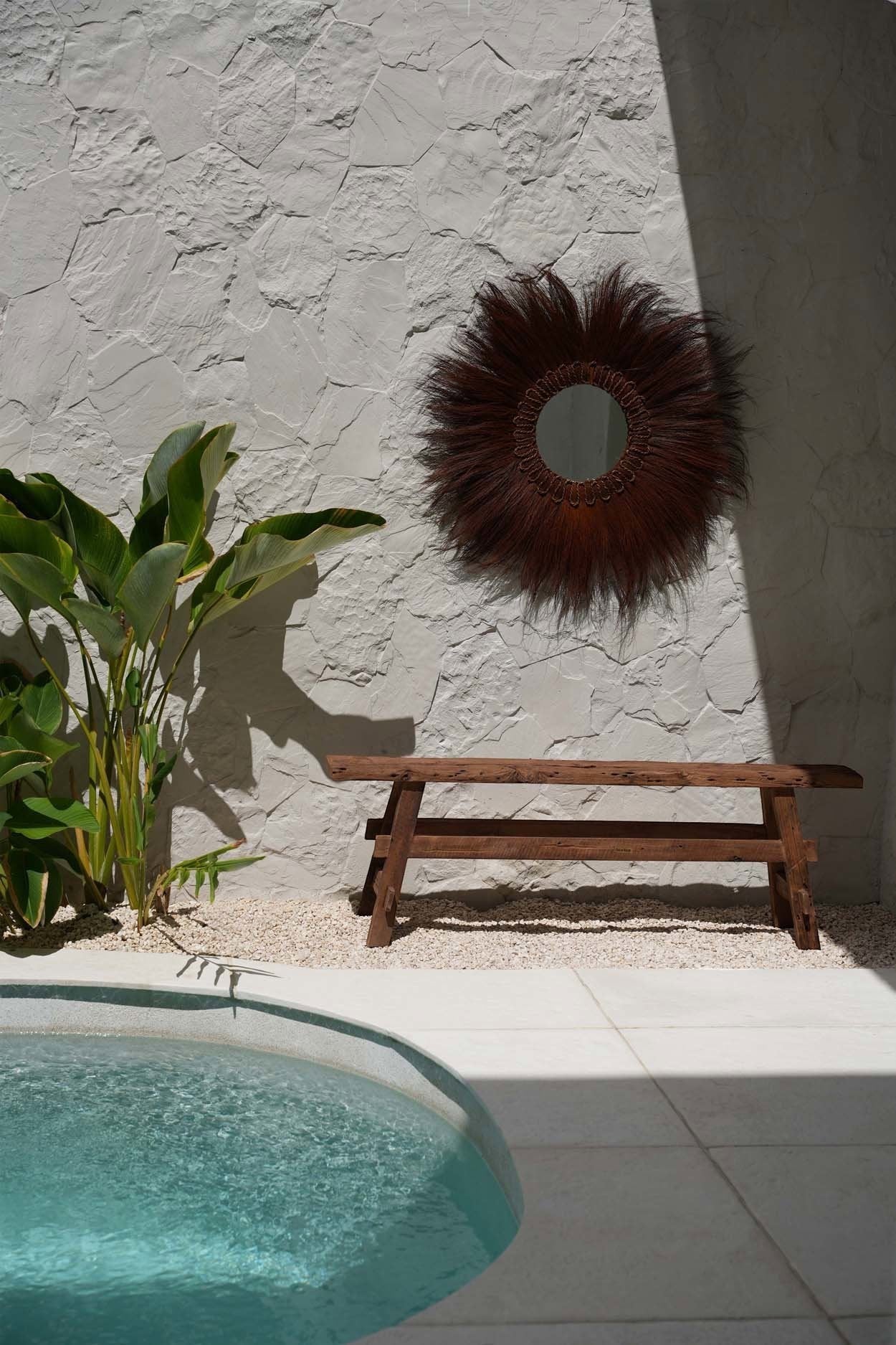 Kayu Living handcrafted solid Indonesian teak rustic bench with natural wood grain finish by a pool and stone wall.