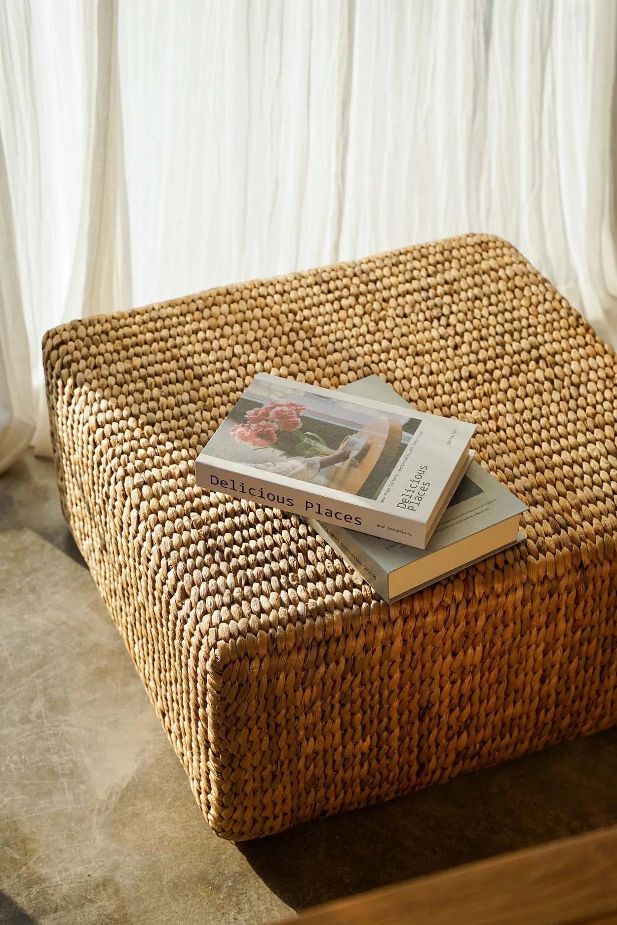 Kayu Living square water-hyacinth pouf in natural tan with a textured woven design, topped with two coffee table books.
