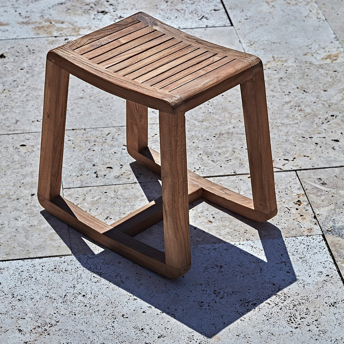 Kayu Living handcrafted solid teak rocking stool with a slatted seat and natural wood grain on a stone patio.