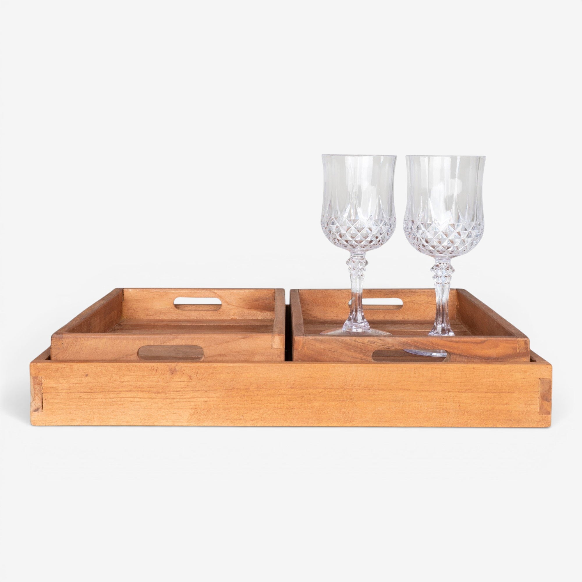 Kayu Living set of 3 natural teak serving trays with cut-out handles. Two crystal wine glasses sit on the largest tray.