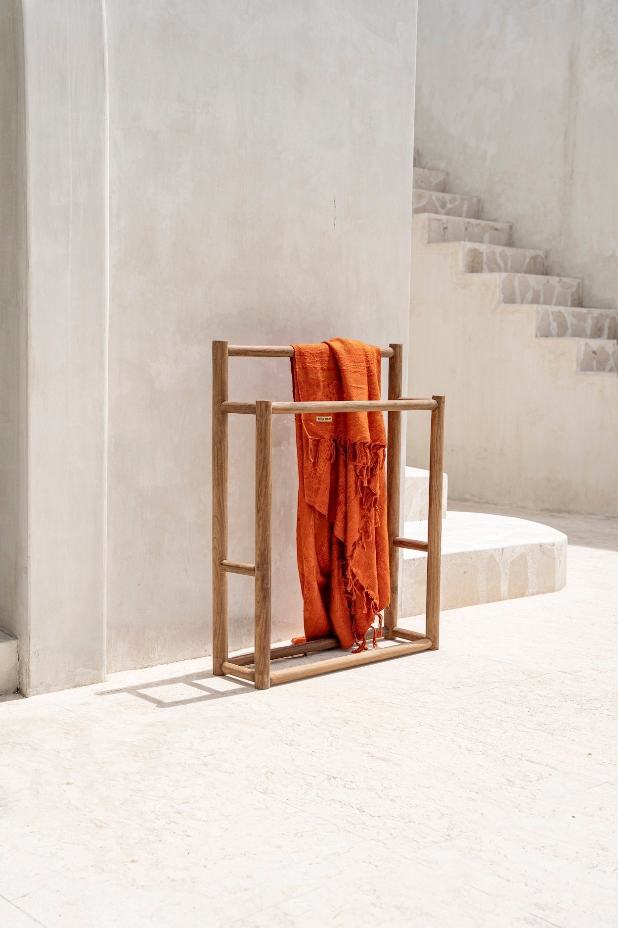 Kayu Living handmade natural teak towel hanger with a draped burnt orange towel in a minimalist outdoor setting.