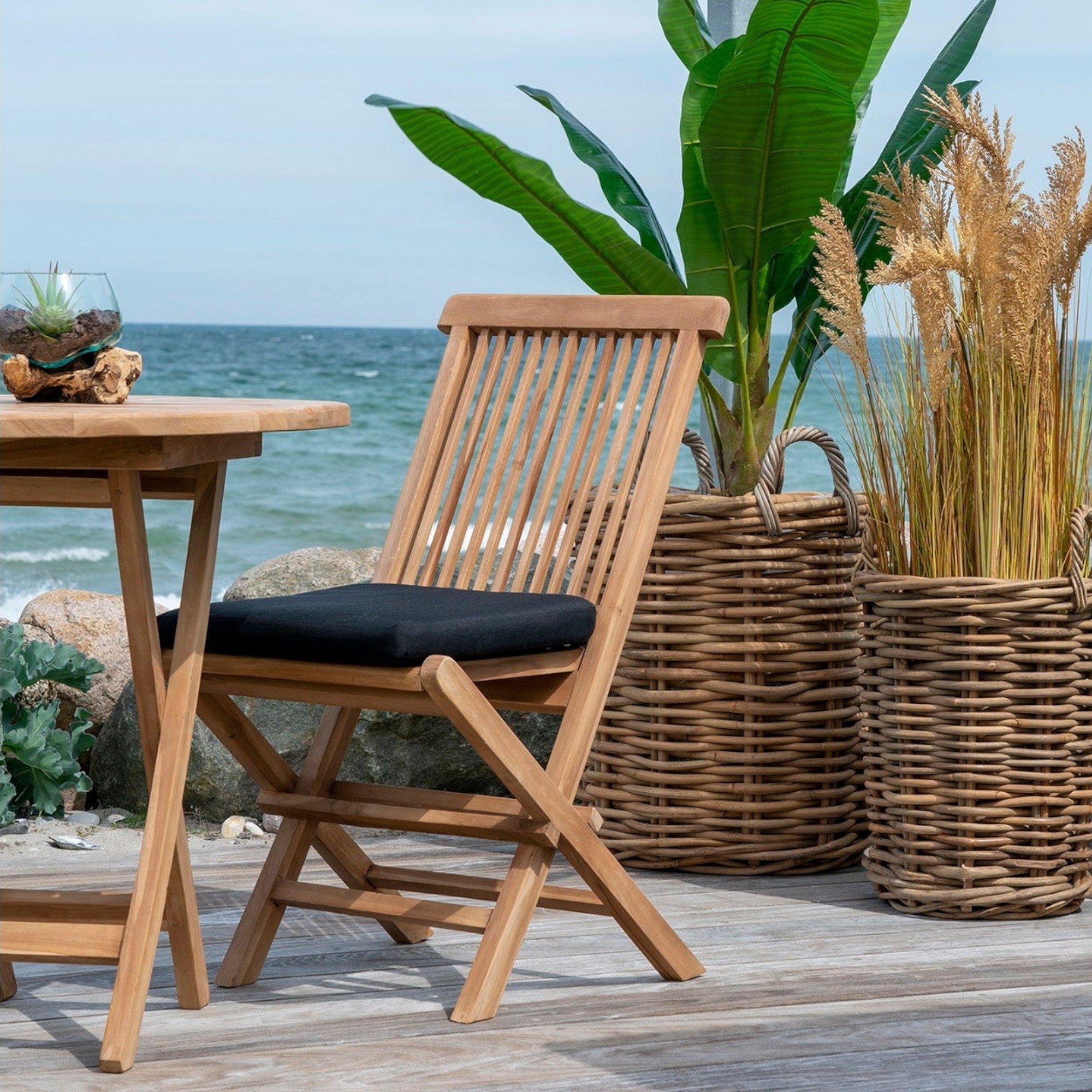 Kayu Living Toledo natural teak wood dining chair with a black cushion, set on a coastal deck by the ocean.