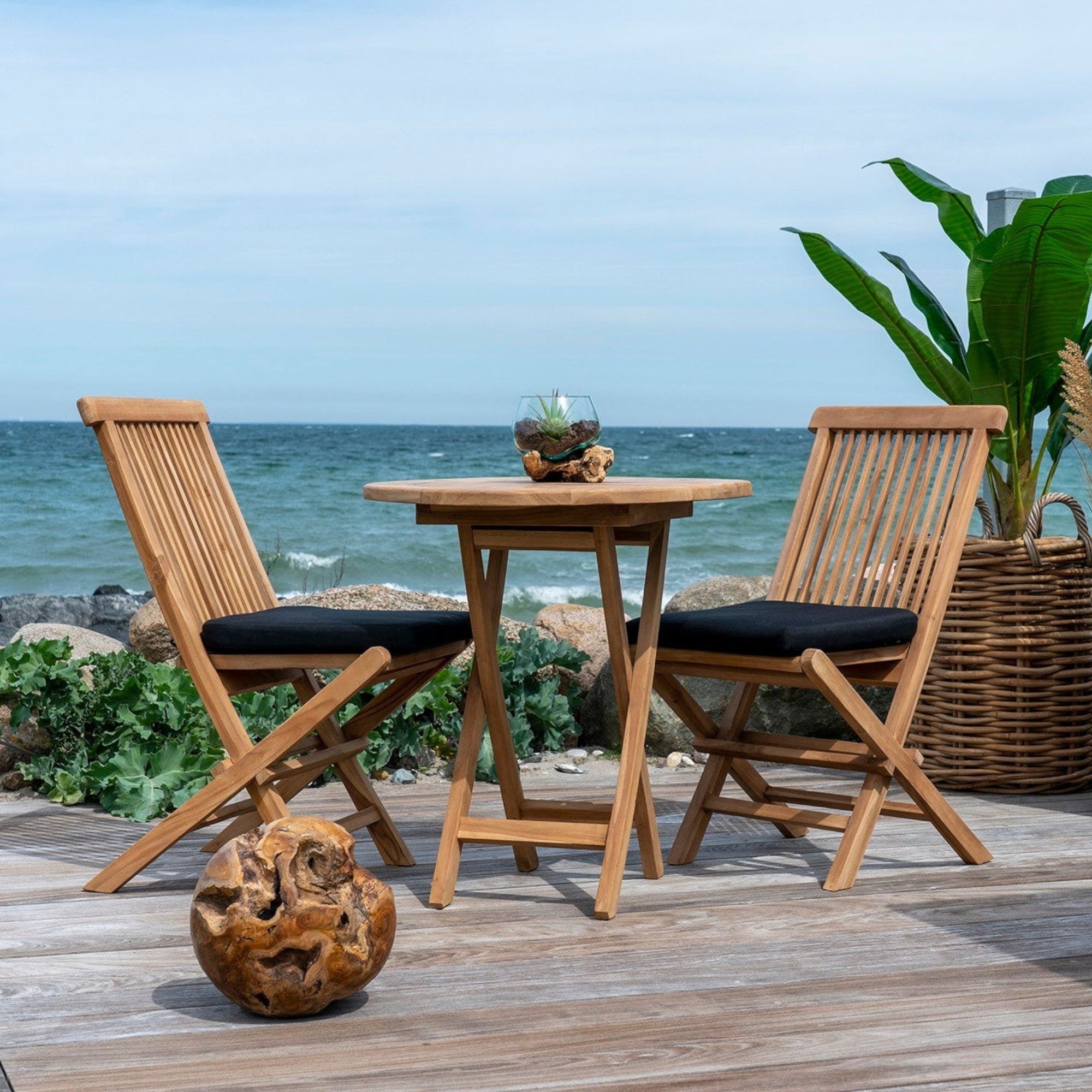 Kayu Living Toledo natural teak dining chairs with black cushions on a coastal patio overlooking the ocean.