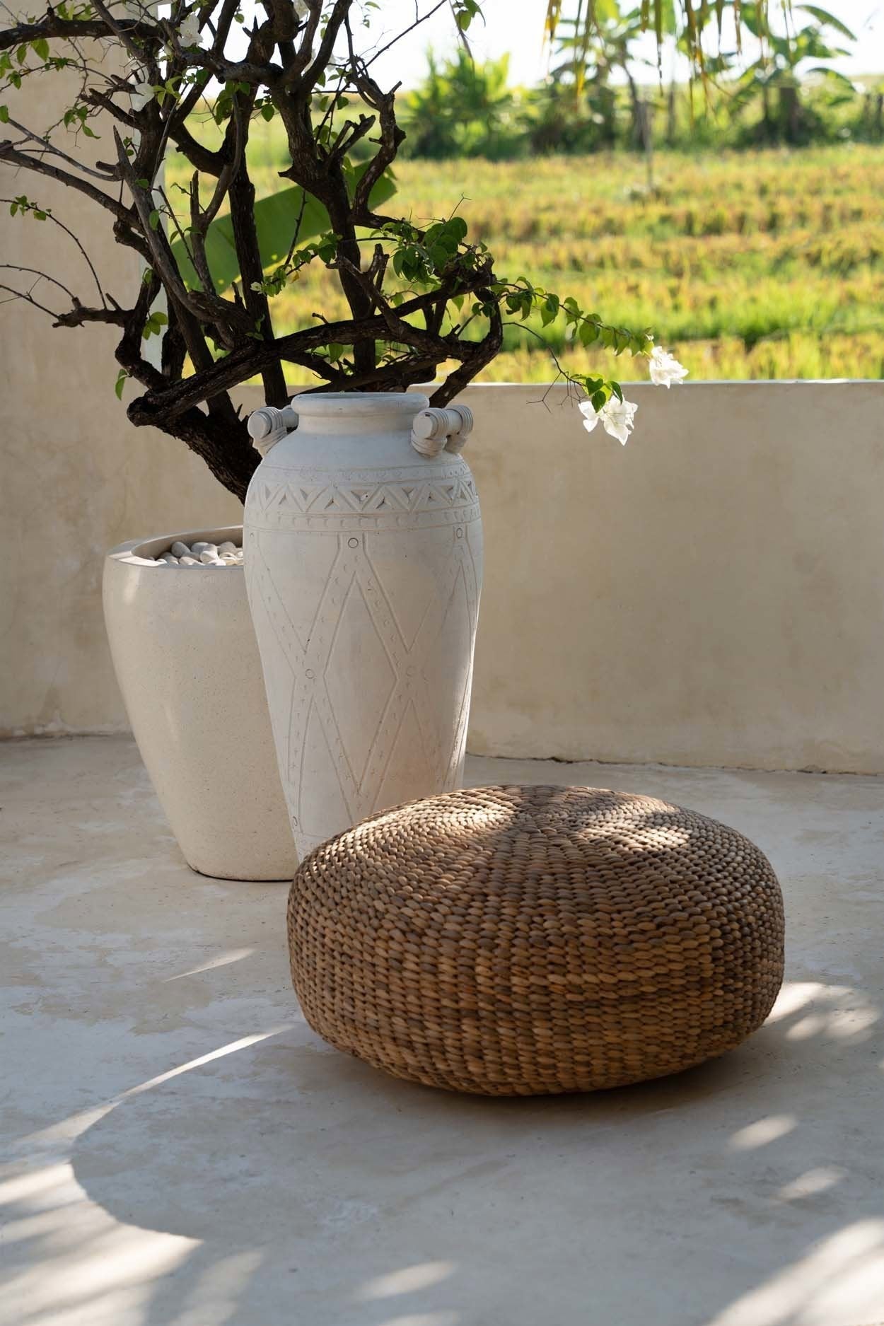 Kayu Living natural water hyacinth pouf on a patio next to a decorative white ceramic vase and flowering tree.