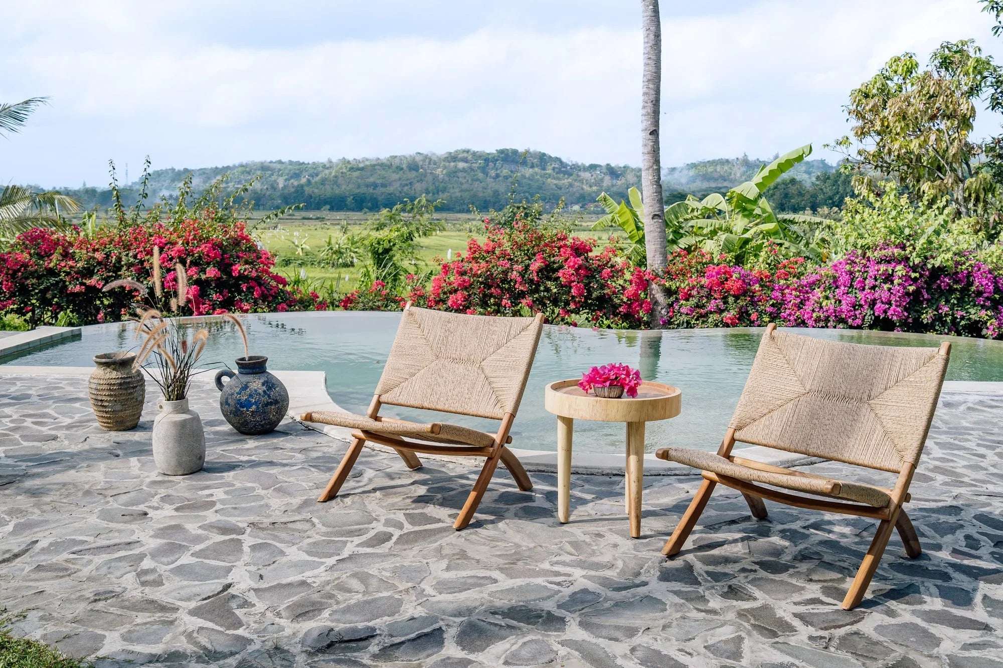 Handmade woven lounge chairs and wooden table by Kayu Living beside pool, with tropical flowers and greenery.