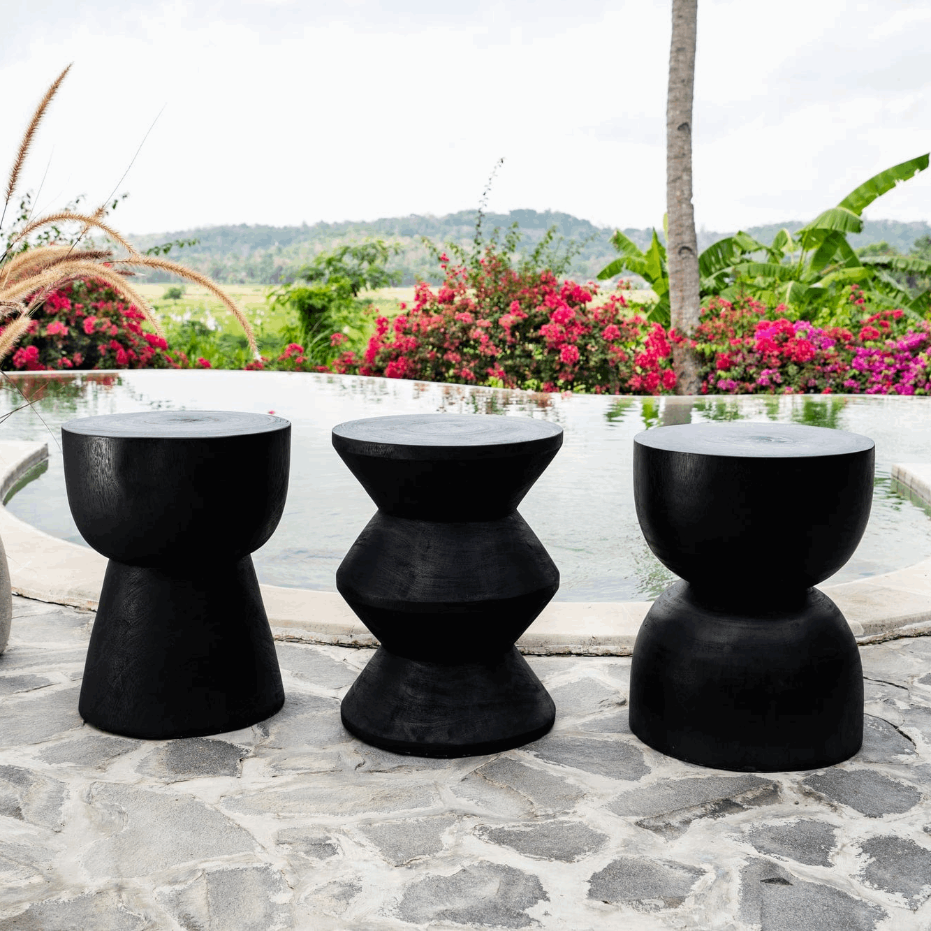Three black hand-carved wooden stools by Kayu Living, poolside with tropical garden view