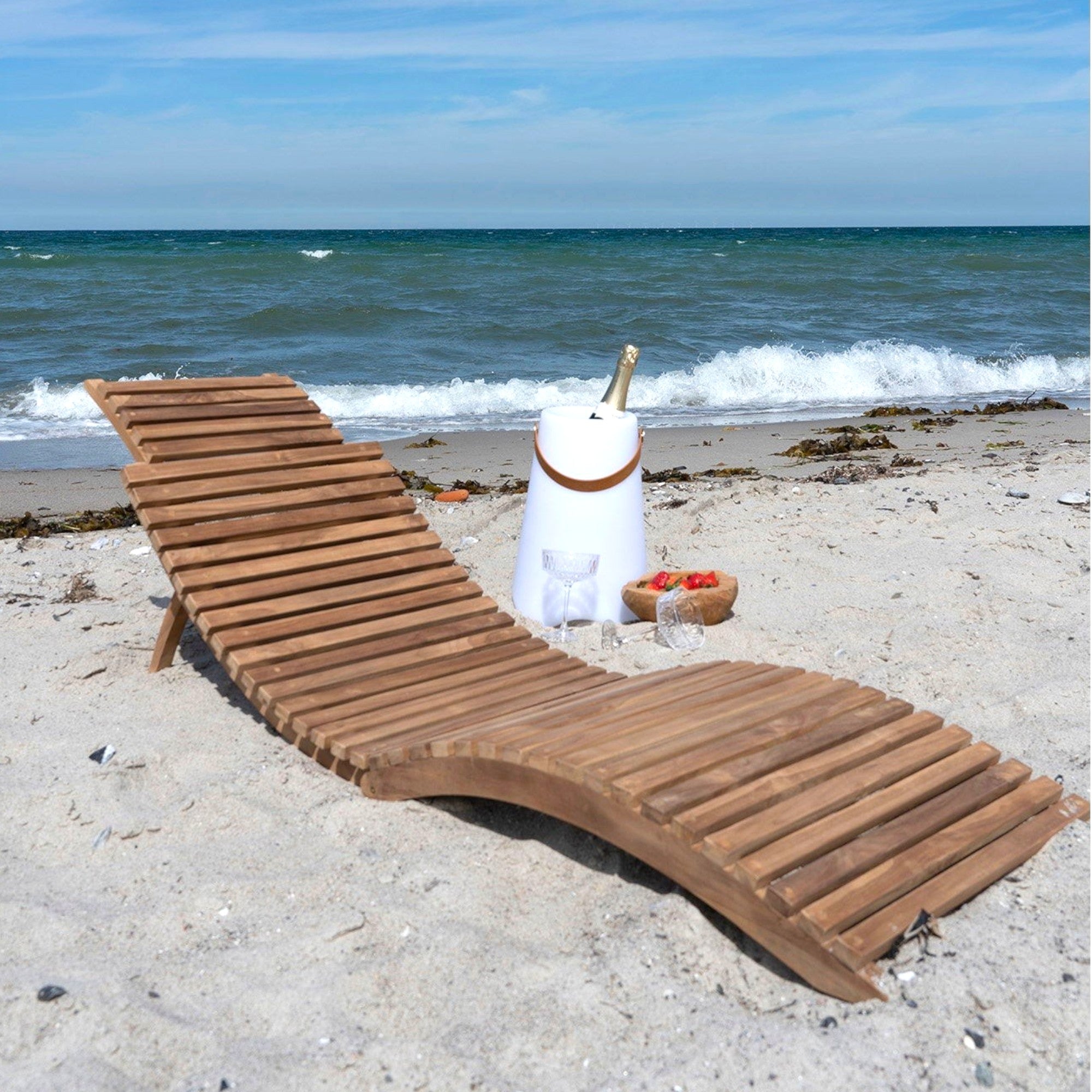 Kayu Living Arrieta natural teak sun lounger on a beach with champagne and strawberries.