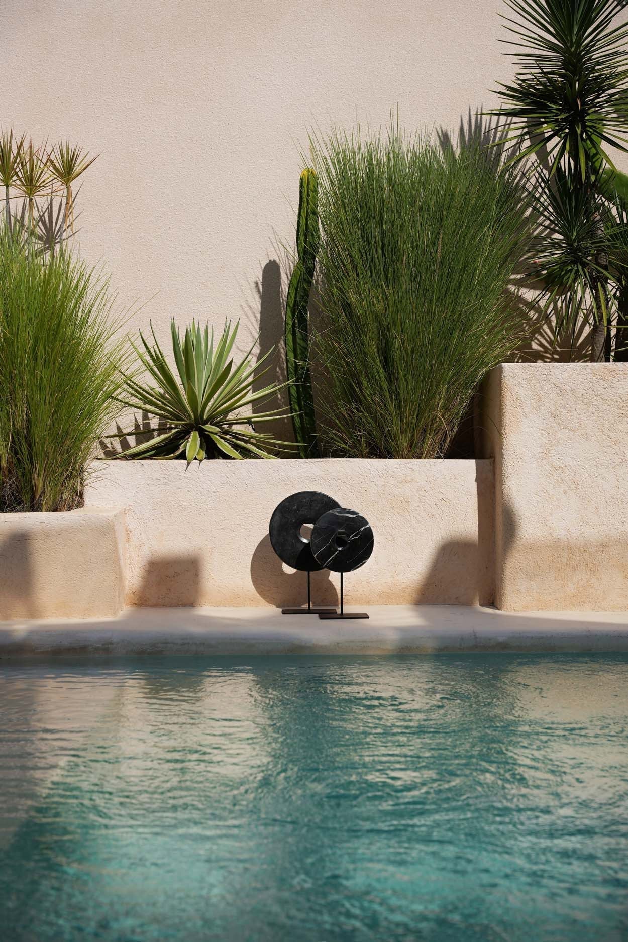 Kayu Living medium black marble disc sculpture with white veining on a metal stand by a pool with desert plants.