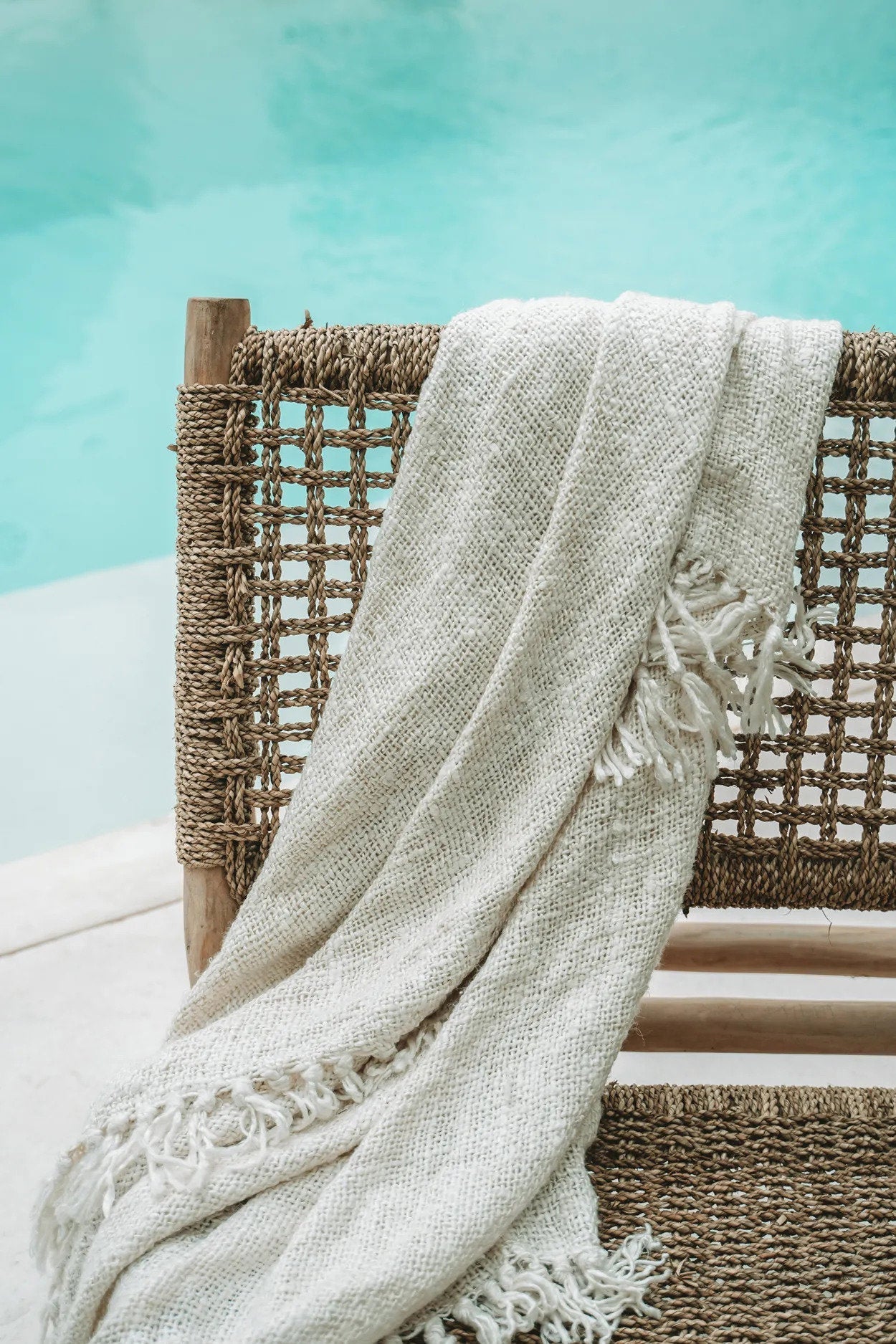 Kayu Living cream hand-woven cotton throw blanket with fringe detail draped over a woven chair by a pool
