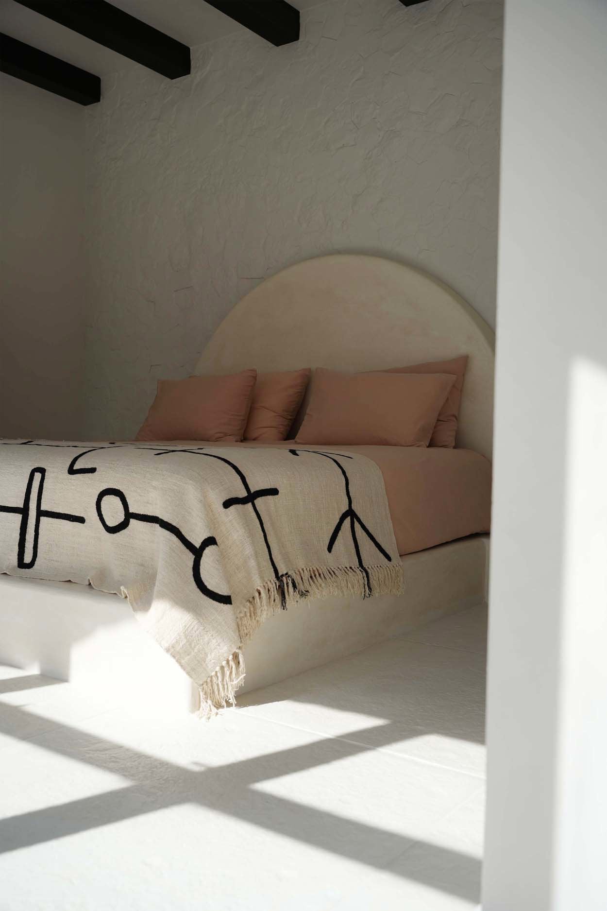 Kayu Living natural black cotton bed throw with evil eye pattern and fringe detail draped over a bed in a bright room.