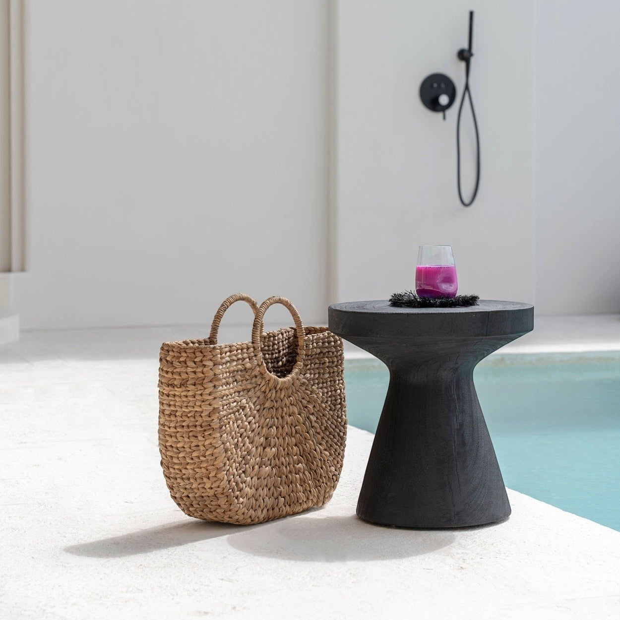 Kayu Living Gravity Side Table in black suar wood, a modern pedestal design with a smooth finish, next to a woven bag by a pool.