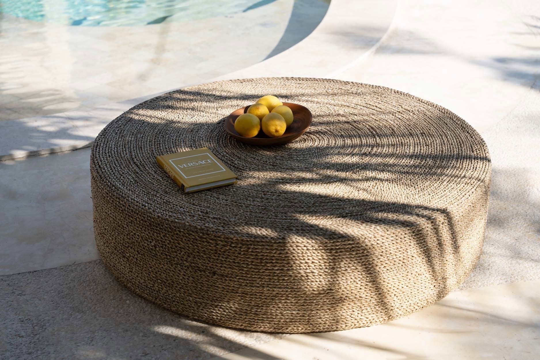 Kayu Living large round seagrass pouf with woven texture, styled as a low coffee table by a pool.