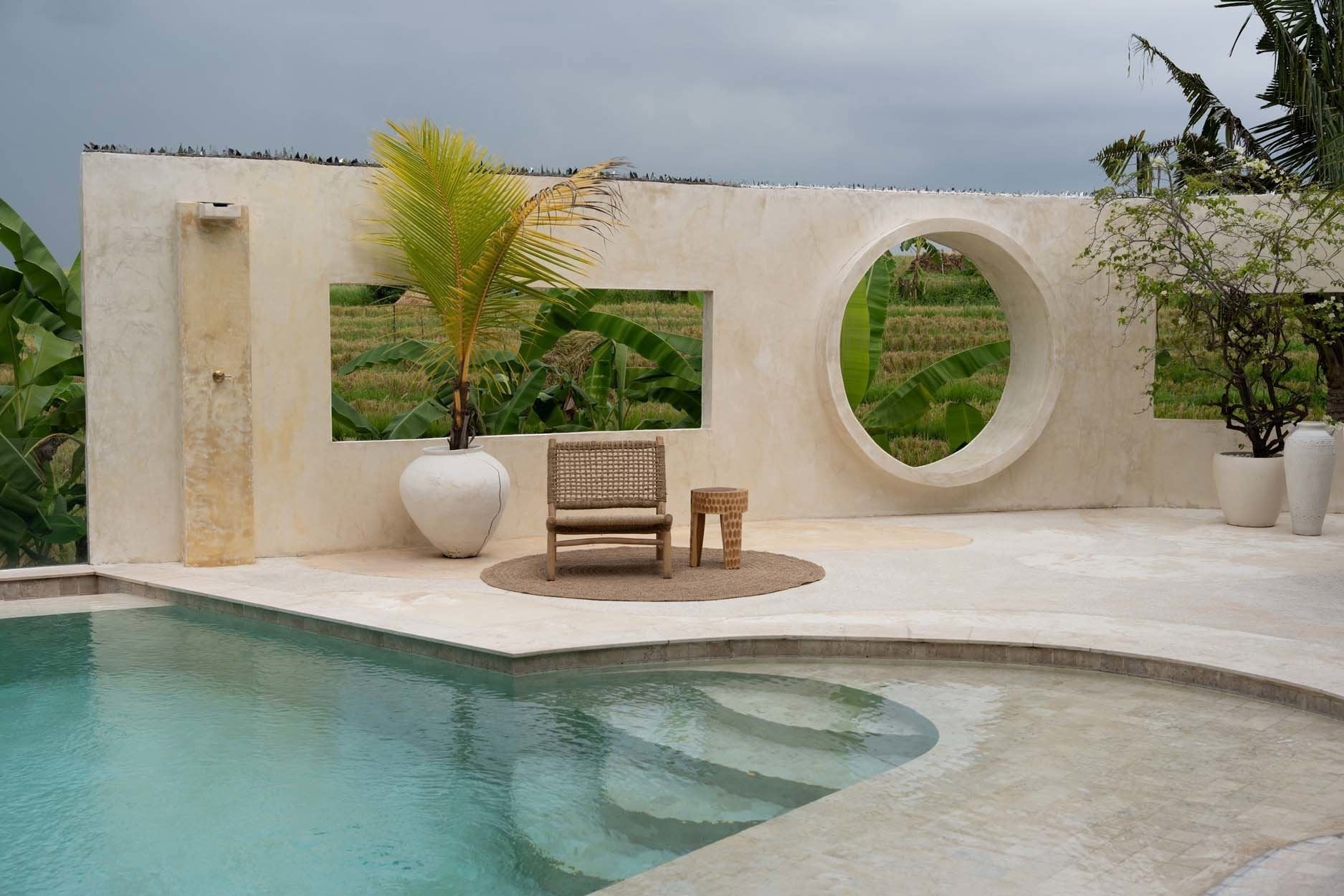 Kayu Living Paramo Stool made of natural suar wood with three legs, placed on a patio by a pool.