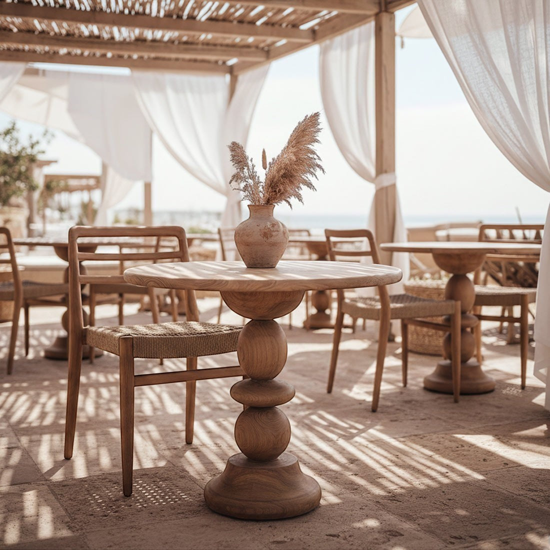 Kayu Living round teak bistro table with a sculptural pedestal base, styled on a sunny outdoor terrace with a vase.