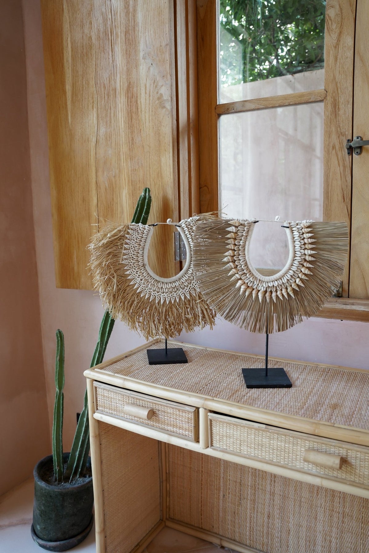 Kayu Living handcrafted seagrass and seashell wall hangers on black stands displayed on a rattan console table.