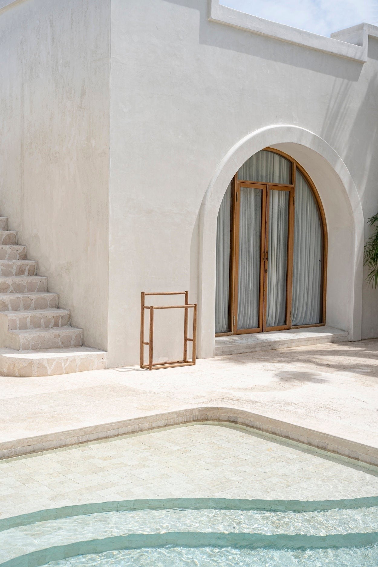 Kayu Living handmade natural teak towel hanger standing on a stone patio by a swimming pool and arched doorway.
